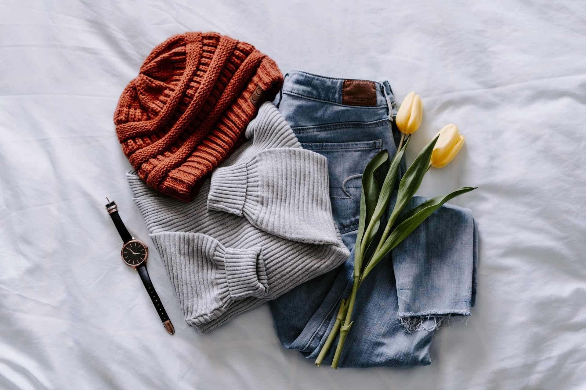 orange-knit-beanie-grey-jumper-blue-pair-of-jeans-and-watch-on-white-bed-travel-capsule-wardrobe